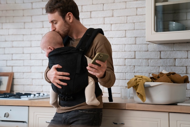 Padre de tiro medio sosteniendo al bebé y haciendo las tareas del hogar