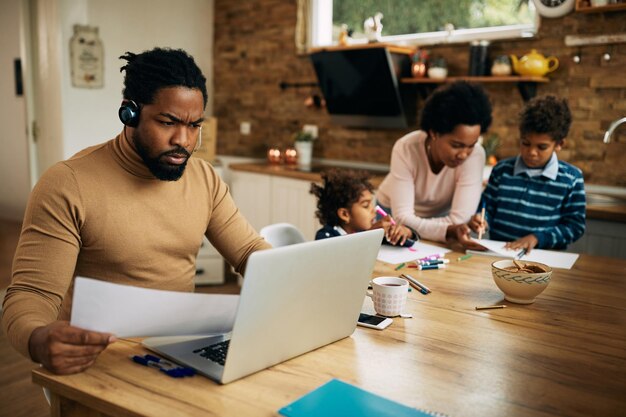 Padre trabajador afroamericano que usa una computadora portátil mientras la madre educa a sus hijos en casa