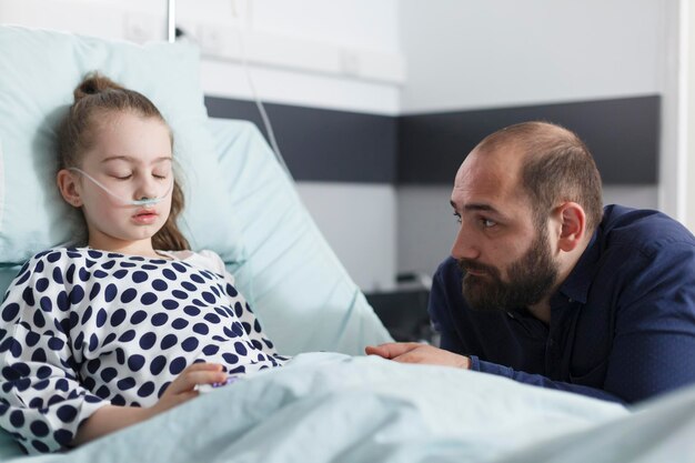 Un padre triste y pensativo mirando a una hijita enferma dormida mientras estaba en la sala de pacientes de la clínica pediátrica. Niña enferma descansando después de un procedimiento médico mientras un padre molesto y preocupado se sienta a su lado.