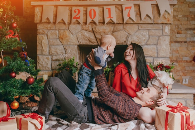 Foto gratuita padre tumbado en el suelo con una chimenea de fondo mientras levanta a su bebé en alto y la madre los mira sonriendo