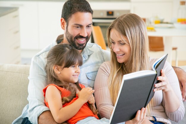 Padres alegres y niña de pelo negro sentados en el sofá en la sala de estar, leyendo un libro juntos y riendo.