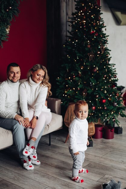 Padres amorosos vestidos de blanco sentados en el sofá cerca del árbol de Navidad mientras miran a su hijo