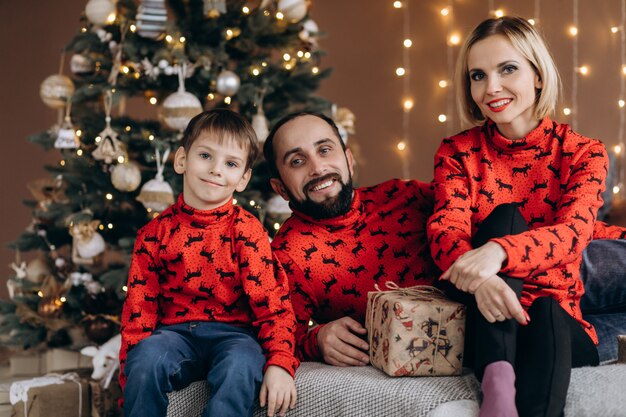 Los padres atractivos y su pequeño hijo en suéteres rojos se divierten abriendo regalos antes de Navidad