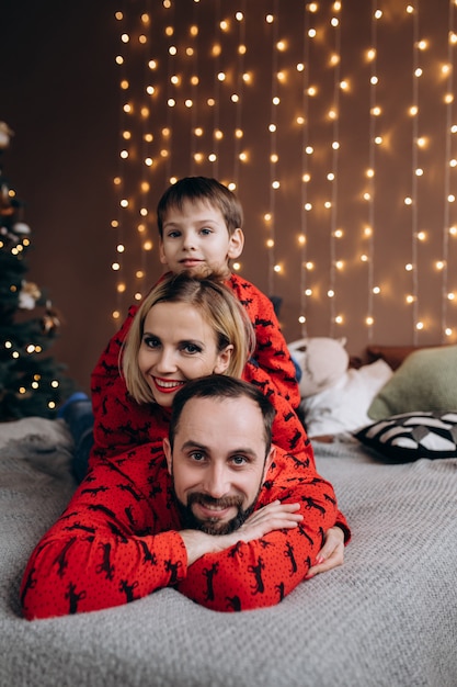 Los padres atractivos y su pequeño hijo en suéteres rojos se divierten descansando en la cama antes de una Navidad.