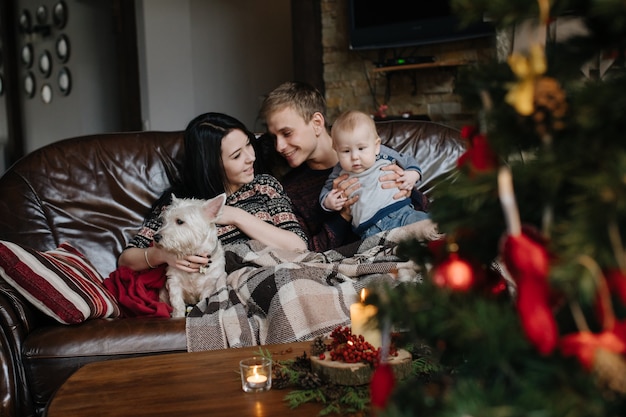 Padres con un bebé en navidad y un perro sentados en el sofá