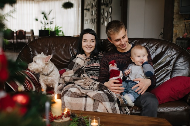 Padres con un bebé en navidad y un perro sentados en el sofá