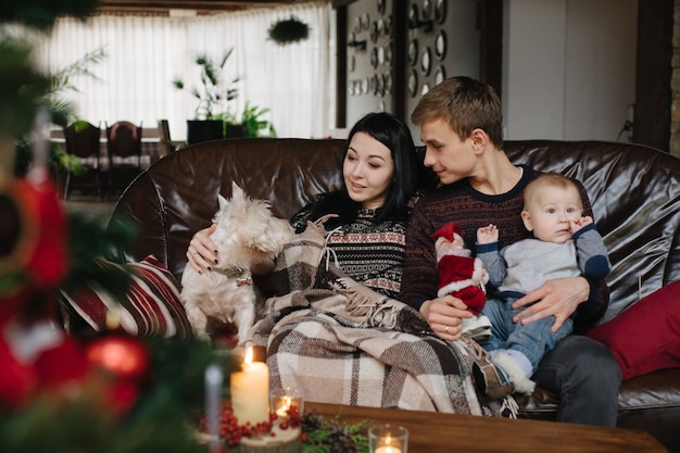 Padres con un bebé en navidad y un perro sentados en el sofá