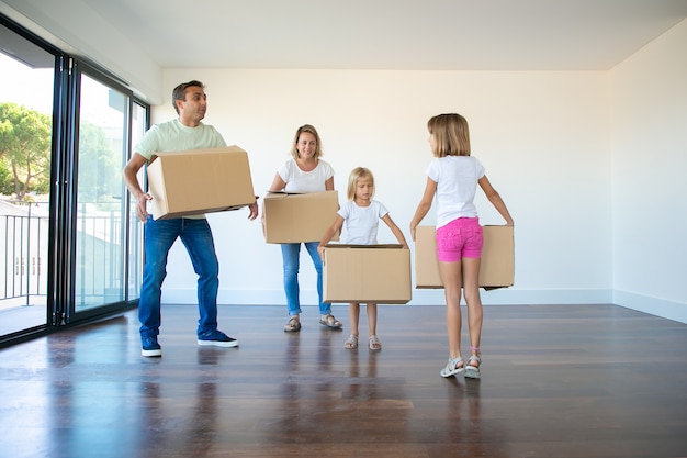 Padres caucásicos y dos niñas sosteniendo cajas de cartón y de pie en la sala de estar vacía