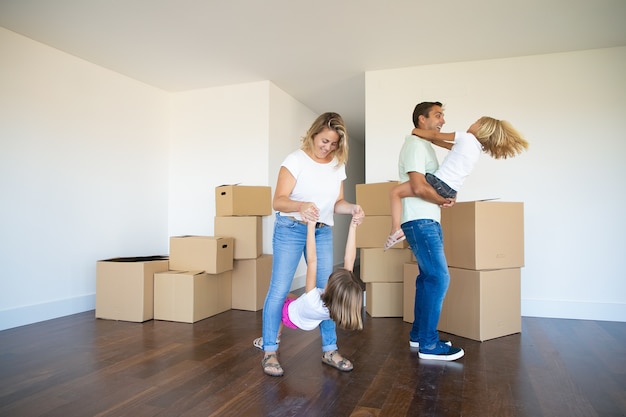 Padres e hijos alegres que disfrutan del nuevo hogar, bailan y se divierten cerca de montones de cajas en una habitación vacía