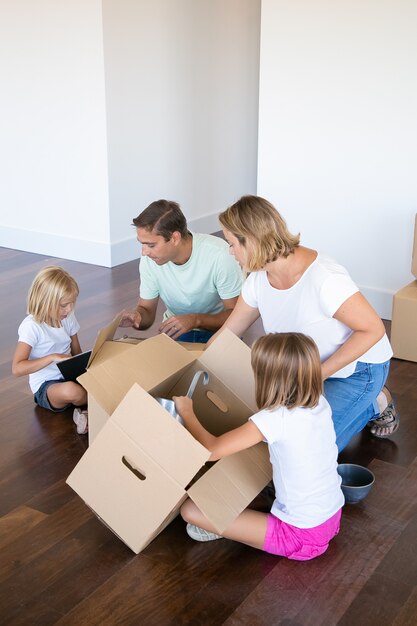 Padres e hijos enfocados desempacando cosas en un apartamento nuevo, sentados en el piso y sacando objetos de cajas abiertas