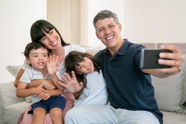 Padres felices abrazando a adorables niños, sentados juntos en el sofá en casa, tomando selfie en el teléfono