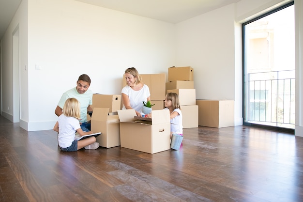 Padres felices y dos niños que se mudan a un nuevo apartamento vacío, sentados en el piso cerca de cajas abiertas