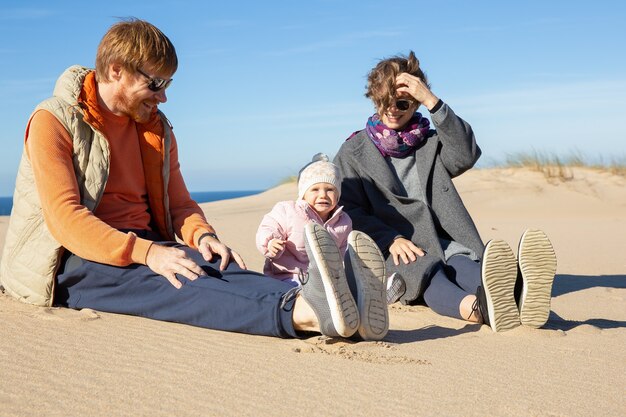 Padres felices y dulce bebé vistiendo ropa abrigada, pasando tiempo libre en el mar, sentados juntos en la arena