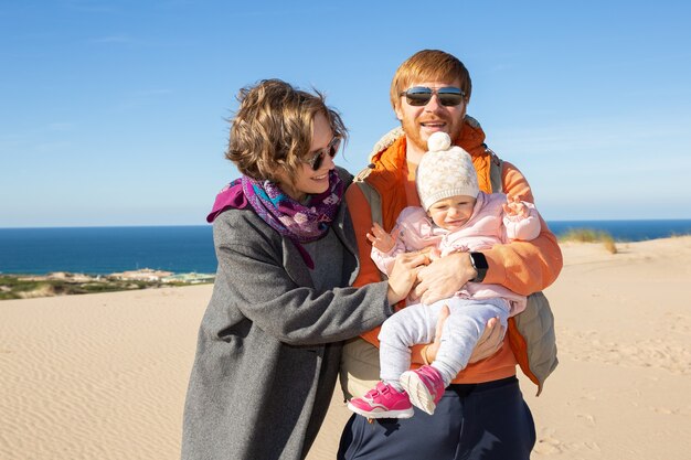 Padres felices con linda hija en brazos mientras está de pie sobre la arena en el mar