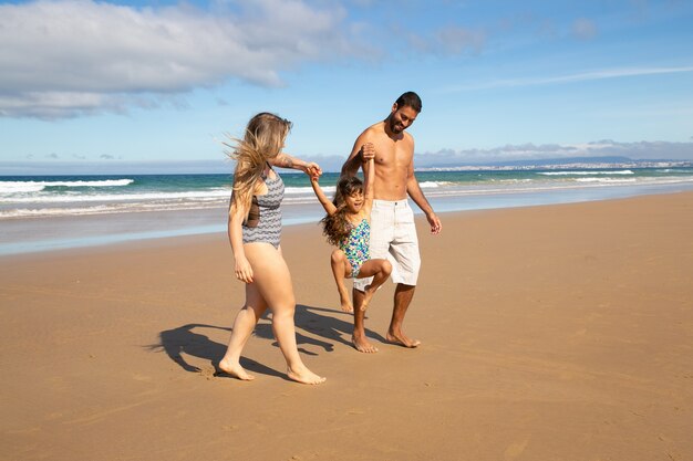 Padres felices y niña vistiendo trajes de baño, caminando sobre arena dorada del mar