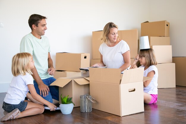 Padres felices y niñas que se divierten mientras desempacan cosas en un apartamento nuevo, se sientan en el piso y toman objetos de cajas abiertas