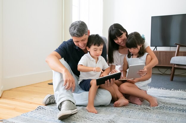 Padres felices y niños lindos que usan dispositivos móviles en el piso de la sala de estar.