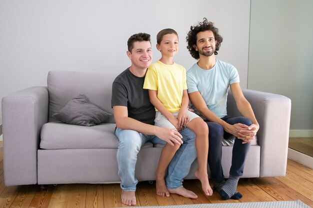 Padres homosexuales felices y niños sentados en el sofá en casa, sonriendo y mirando a otro lado. Copie el espacio. Concepto de familia y paternidad