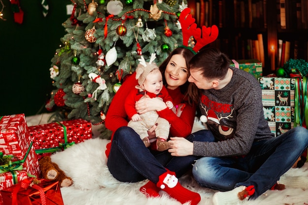 Los padres juegan con la pequeña hija con sombrero de venado que la sostiene en sus brazos y se sienta delante de un árbol de Navidad