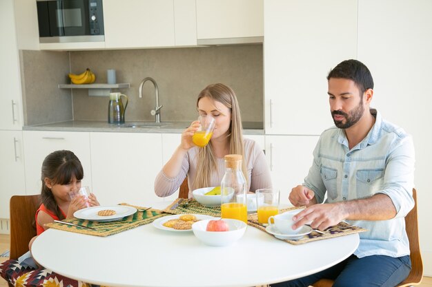 Los padres y el niño desayunando, bebiendo jugo de naranja, sentados en la mesa de comedor con frutas y galletas.