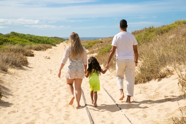 Foto gratuita los padres y el niño vistiendo ropa de verano, caminando por el camino de arena hacia el mar, niña cogidos de la mano de los padres