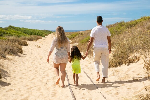 Los padres y el niño vistiendo ropa de verano, caminando por el camino de arena hacia el mar, niña cogidos de la mano de los padres
