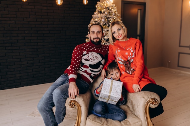 Los padres se sientan alrededor de un niño pequeño en una silla suave ante un árbol de Navidad