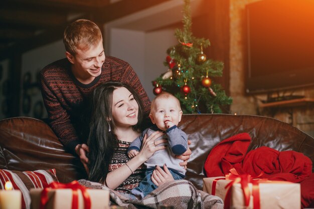 Padres sonriendo mirando a su bebé con la mano en la boca y con regalos de navidad