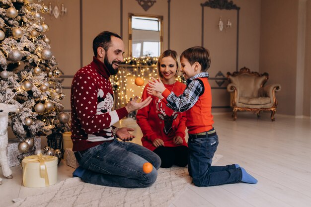 Los padres y su pequeño hijo en suéter rojo se divierten con naranjas sentadas ante un árbol de Navidad