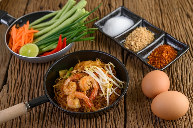 Padthai camarones en un recipiente negro con huevos y condimentos en la mesa de madera.