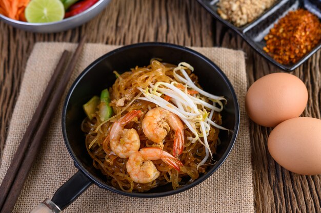 Padthai camarones en un recipiente negro con huevos y condimentos en la mesa de madera.