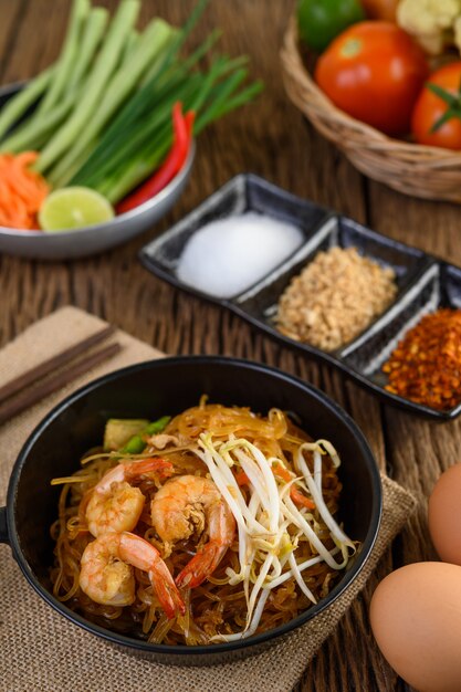 Padthai camarones en un recipiente negro con huevos y condimentos en la mesa de madera.