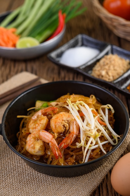 Padthai camarones en un recipiente negro con huevos y condimentos en la mesa de madera.