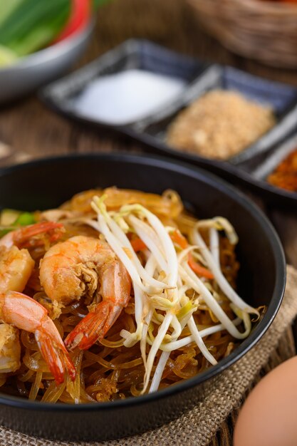 Padthai camarones en un recipiente negro con huevos y condimentos en la mesa de madera.