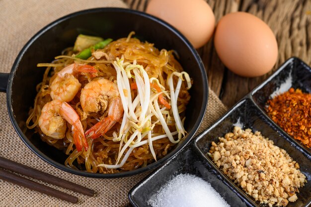 Padthai camarones en un recipiente negro con huevos y condimentos en la mesa de madera.