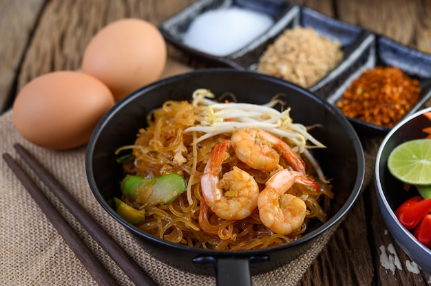 Padthai camarones en un recipiente negro con huevos y condimentos en la mesa de madera.