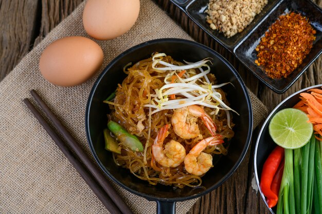 Padthai camarones en un recipiente negro con huevos y condimentos en la mesa de madera.