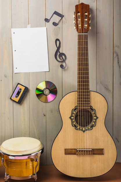 Página musical en blanco; casete; disco compacto; y nota musical pegada en pared de madera con guitarra y bongo drum.