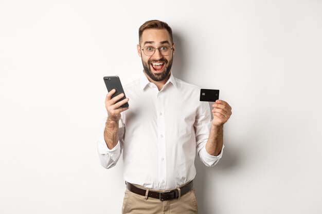 Pago comercial y online. Hombre emocionado pagando con teléfono móvil y tarjeta de crédito, sonriendo asombrado, de pie