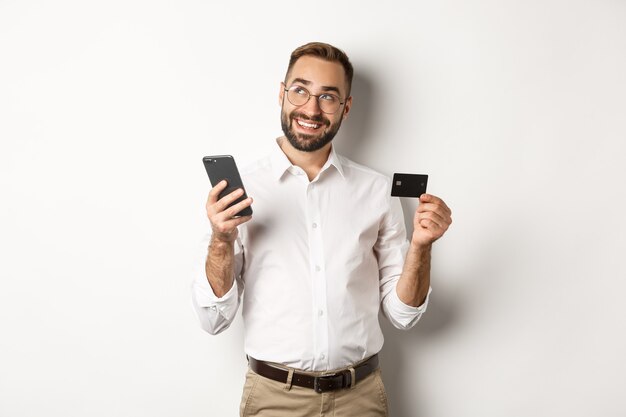 Pago comercial y online. Imagen de hombre guapo pensando mientras sostiene la tarjeta de crédito y el teléfono inteligente, de pie