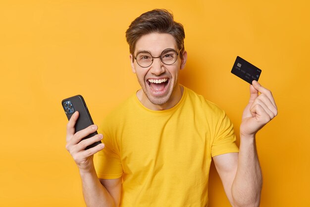 Pagos fáciles y concepto de compras en línea Un hombre alegre usa una camiseta informal sostiene un celular y una tarjeta de crédito exclama de alegría usa gafas redondas una camiseta informal aislada sobre un fondo amarillo