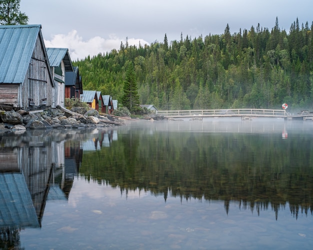 Paisaje de bosque y casas reflexionando sobre un lago cubierto de niebla bajo un cielo nublado