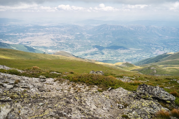 Paisaje de colinas cubiertas de vegetación con montañas rocosas bajo un cielo nublado en el fondo