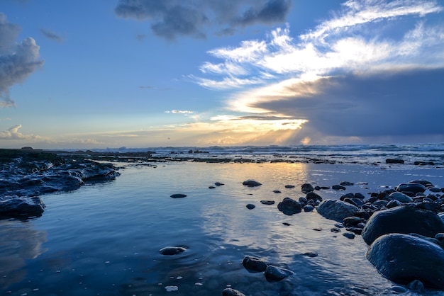 Paisaje de un mar cubierto de rocas bajo la luz del sol y un cielo nublado durante la puesta de sol