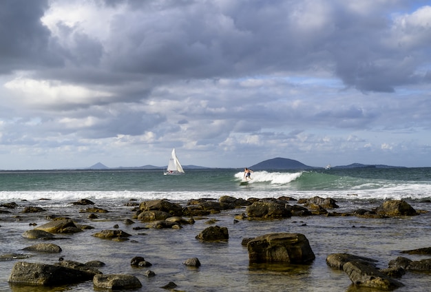 Foto gratuita paisaje de la orilla rodeada por el mar con barcos y surfistas bajo un cielo nublado