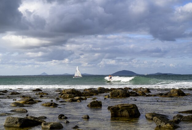 Paisaje de la orilla rodeada por el mar con barcos y surfistas bajo un cielo nublado