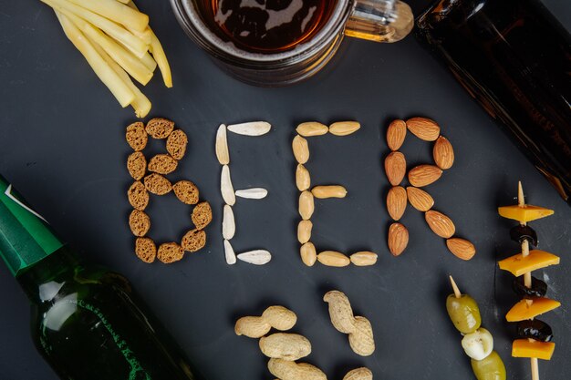 Una palabra cerveza hecha de nueces crujientes saladas, semillas de girasol y galletas saladas, bocadillos salados con botellas de cerveza de queso y una jarra de cerveza en la vista superior negra