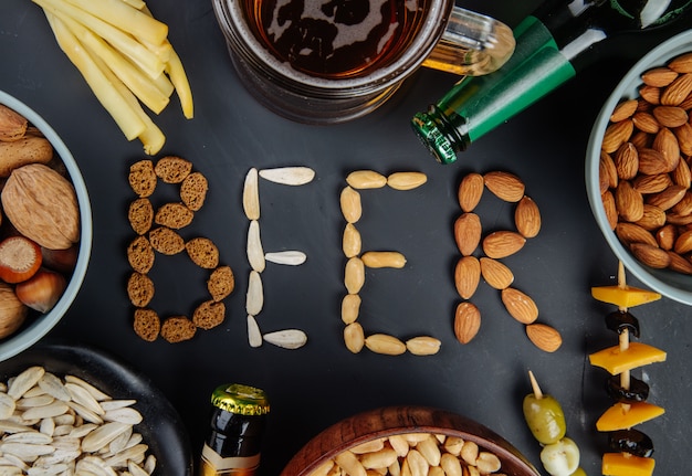 Una palabra cerveza hecha de nueces crujientes saladas, semillas de girasol y galletas saladas, bocadillos salados con botellas de cerveza de queso y una jarra de cerveza en la vista superior negra