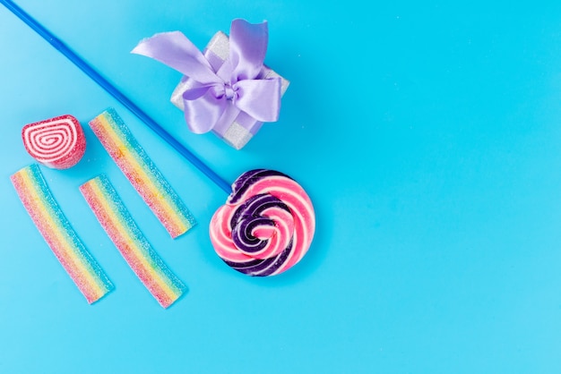 Una paleta dulce de vista superior con palo azul y cajita de regalo púrpura en el escritorio azul, cumpleaños de azúcar dulce