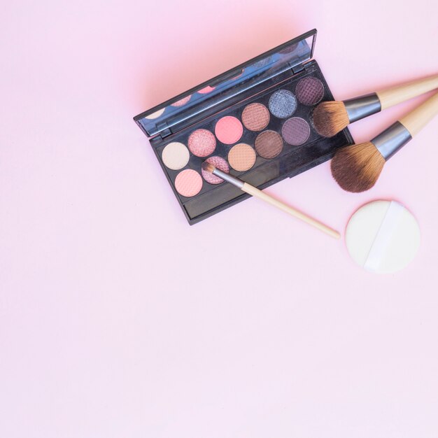 Una paleta de sombras de ojos abierta con pincel de maquillaje y esponja sobre fondo rosa